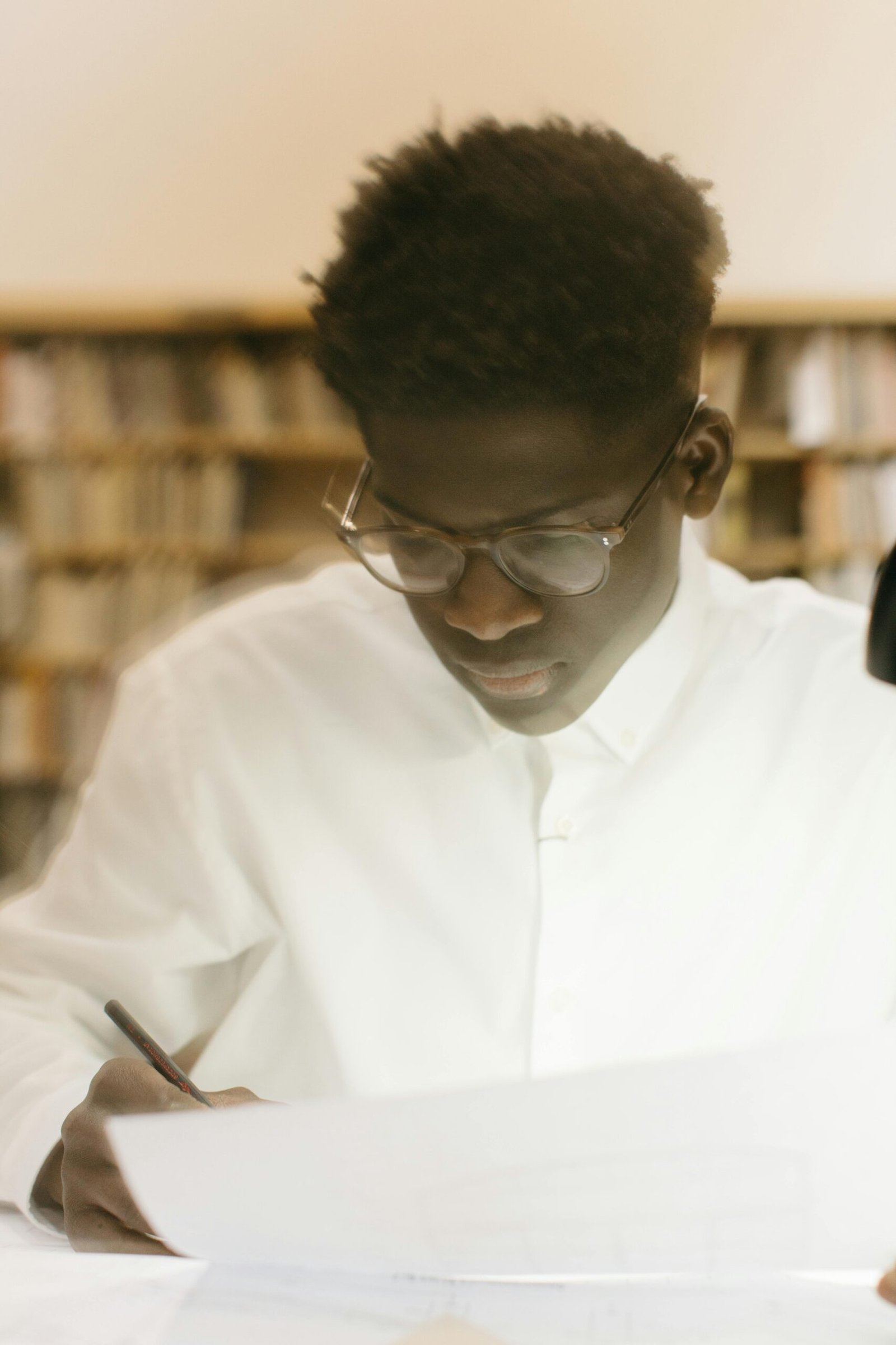 Young man with glasses writing in a library, deeply focused on his work.